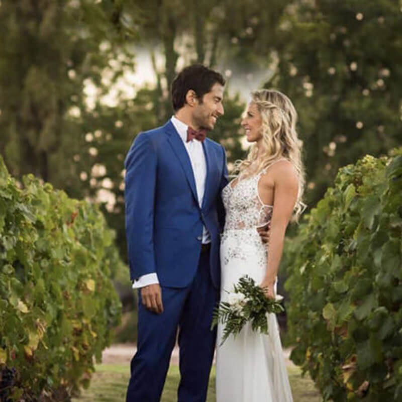 Fotografia de um homem e uma mulher vestidos de noivos, abraçados, olhando nos olhos um do outro, em pé em um jardim em plena luz do dia.